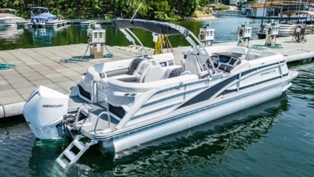 A white and blue Bennington 25 pontoon boat with a 15-person capacity in the water at a marine fuel dock.