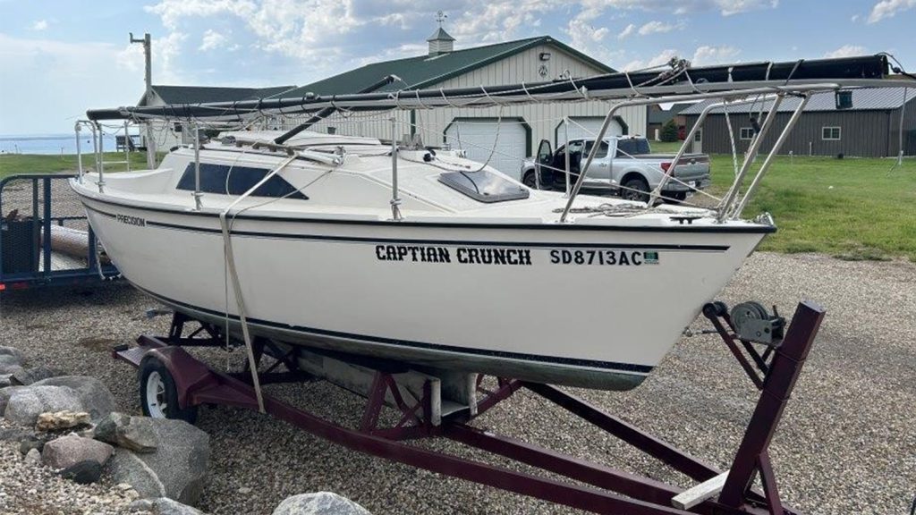 A small white sailboat capable of carrying two to four people strapped to a boat trailer with its sails down.